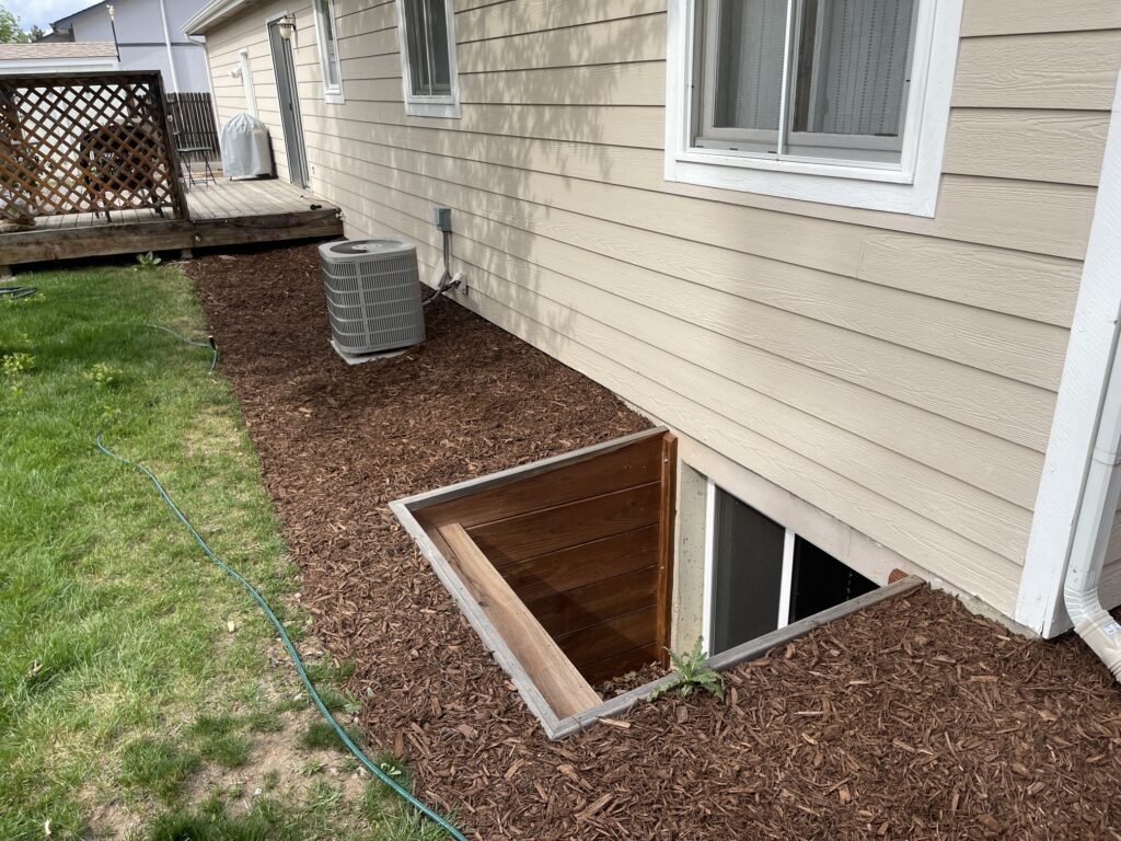 Custom wood egress window well surrounded by fresh mulch installation in Windsor Colorado.