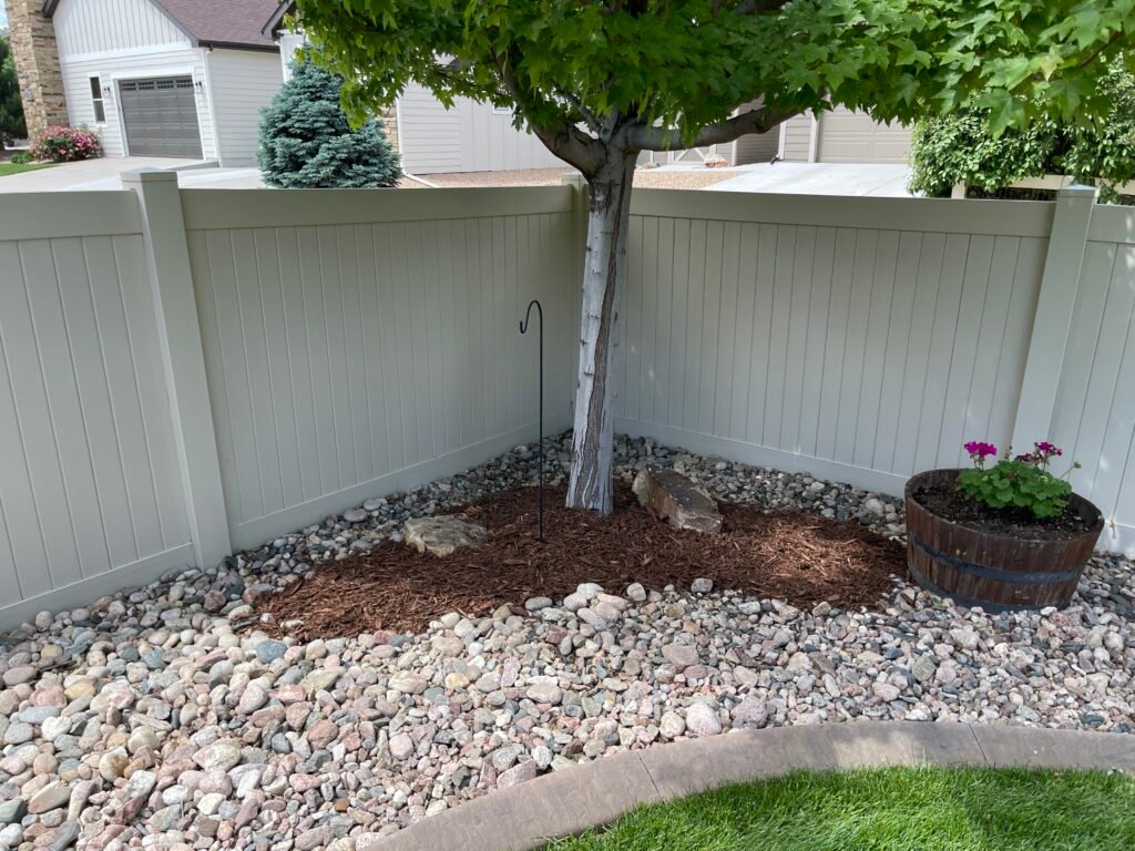Vinyl privacy fence corner with decorative river rock, a fresh mulch ring beneath a pruned tree, and a barrel planter with small flowers in a Johnstown, Colorado backyard.