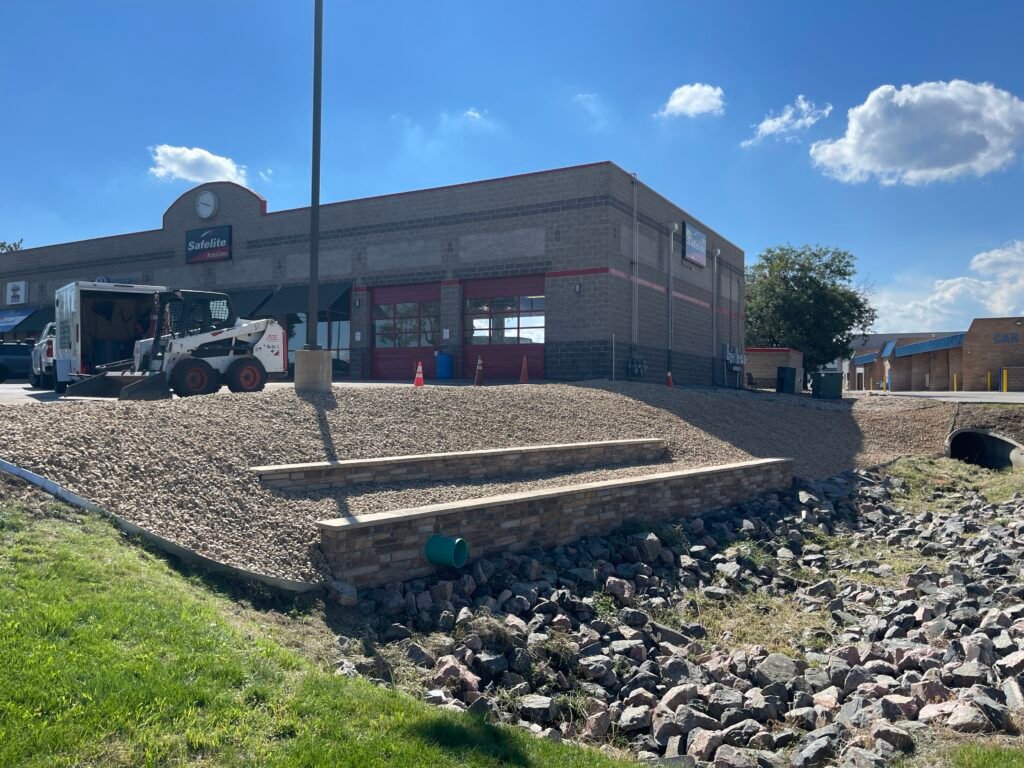 Landscape construction crew using a skid steer loader to install a stacked stone retaining wall and grade rock slope in Northern Colorado.
